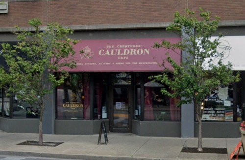 Exterior of "The Creatures Cauldron Café," featuring a maroon awning and trees flanking the entrance.