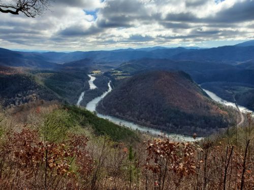 A scenic view of a winding river surrounded by mountains and autumn foliage under a cloudy sky.