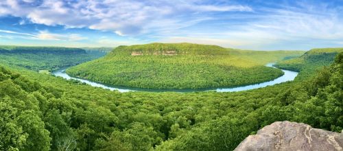 A panoramic view of a lush green valley with a winding river and a distant plateau under a blue sky.