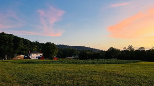 A serene landscape featuring a white house, green fields, and a colorful sunset over rolling hills.