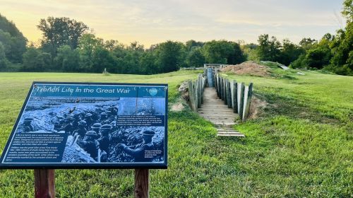 A sign about trench life in WWI next to a reconstructed trench in a grassy area, with trees in the background.