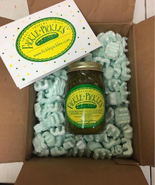 A jar of pickles in a cardboard box filled with packing peanuts, alongside a card from Fickle Pickles.