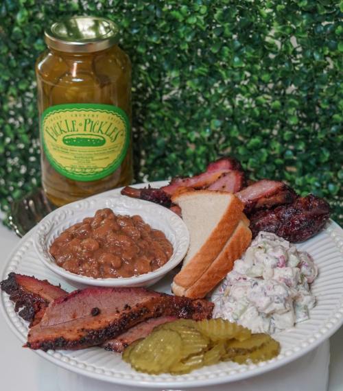 A plate of barbecue with brisket, pickles, coleslaw, baked beans, and a slice of bread, alongside a jar of pickles.