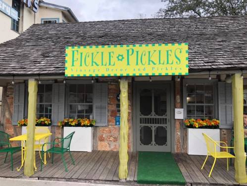 A colorful storefront with a sign reading "Fickle Pickles" and flower boxes, featuring vintage decor and pickles.