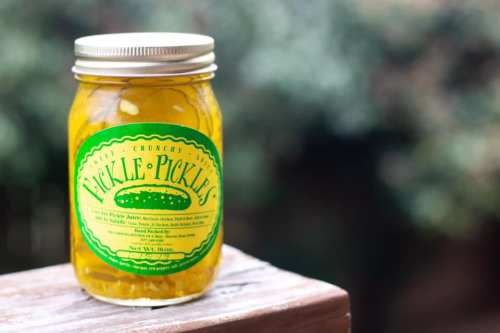 A jar of pickles with a bright green label, sitting on a wooden surface, against a blurred outdoor background.