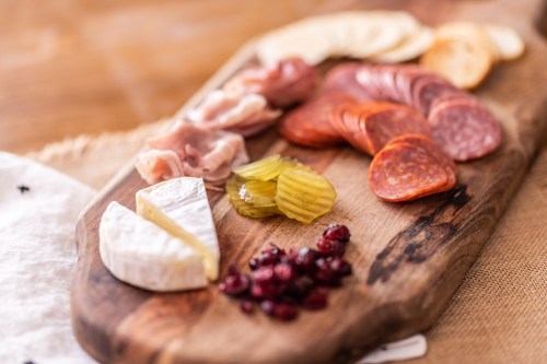 A wooden charcuterie board featuring assorted meats, cheeses, pickles, and dried cranberries.