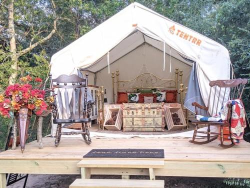 A cozy glamping tent with a bed, decorative pillows, rocking chairs, and flowers on a wooden deck in a natural setting.