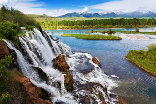 A scenic waterfall cascades into a river, surrounded by lush greenery and mountains under a clear blue sky.