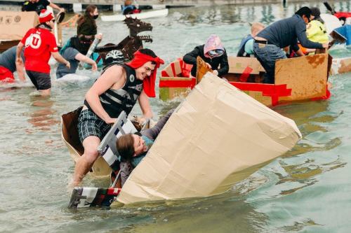 Participants in cardboard boats race in a water event, some wearing costumes and enjoying the fun.