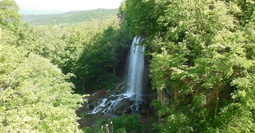 A serene waterfall cascading down rocky cliffs, surrounded by lush green trees and a mountainous landscape.