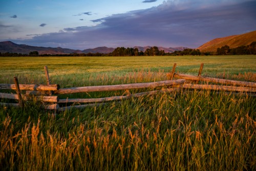 A rustic wooden fence in a grassy field, with mountains and a colorful sky in the background during sunset.
