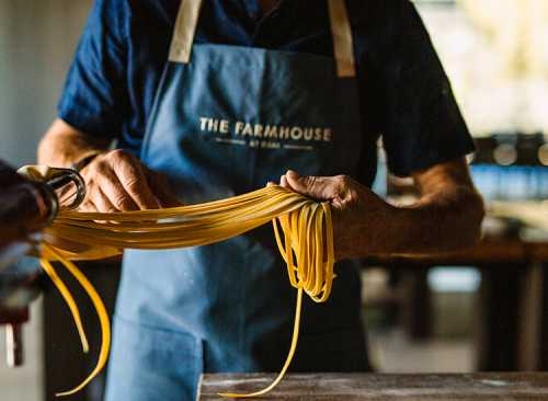 A person in an apron holds freshly made pasta strands in a kitchen setting.