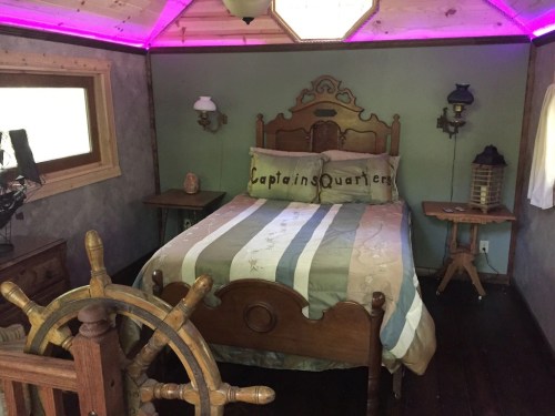 Cozy bedroom with a nautical theme, featuring a bed with "Captain's Quarters" pillow and ship wheel decor.