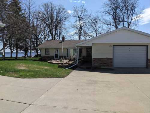 A single-story house with a garage, surrounded by green grass and trees, near a body of water under a clear blue sky.