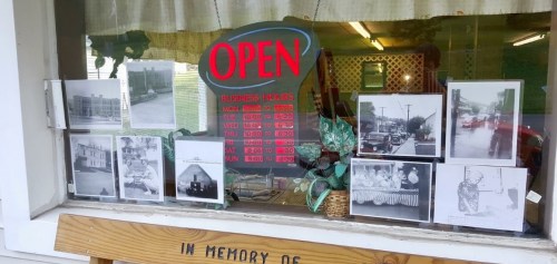 A shop window displays an "OPEN" sign and historical black-and-white photos, with business hours listed above.