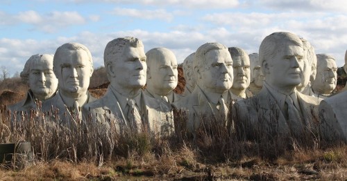 A row of large, weathered stone busts of various historical figures in a grassy field under a cloudy sky.