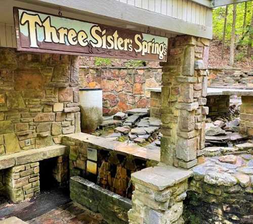 Sign for "Three Sisters Springs" above a stone structure with flowing water and a natural setting.