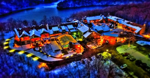 Aerial view of a cozy lodge surrounded by snow, with warm lights glowing against a backdrop of trees and a river.