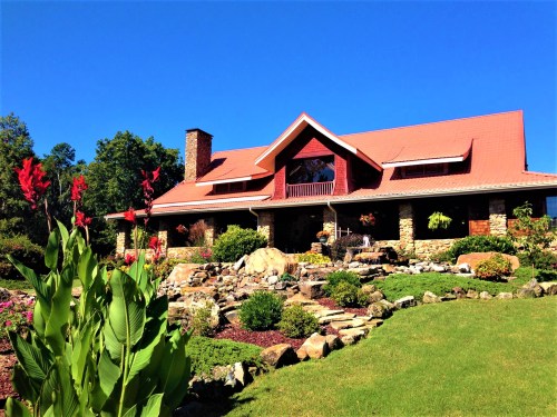 A large, picturesque house with a red roof, surrounded by lush greenery and colorful flowers under a clear blue sky.