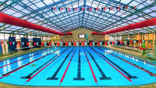 Indoor swimming pool with clear blue water, marked lanes, and colorful flags hanging from the ceiling.