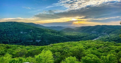 A scenic view of lush green mountains under a cloudy sky, with the sun setting in the background.