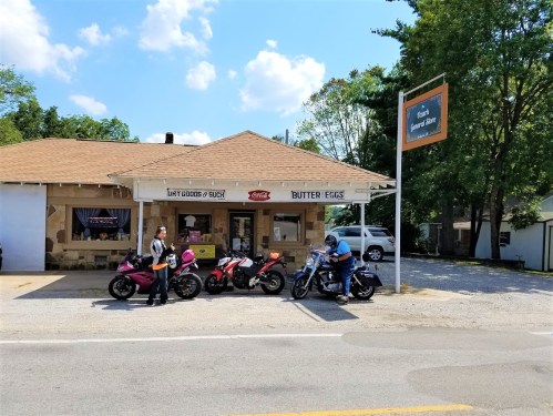 Two motorcyclists stand beside their bikes in front of a small store with a sign reading "Butter Eggs." Sunny day.