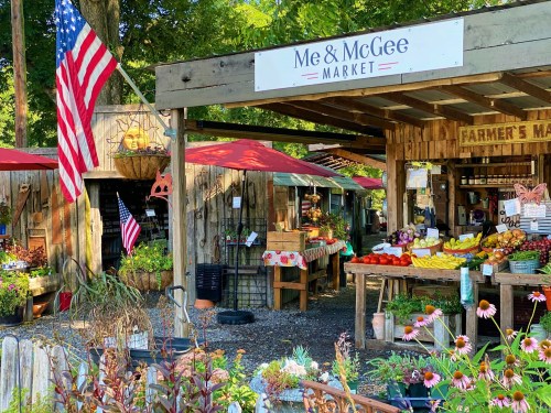 A charming market with wooden stalls, colorful produce, and American flags, surrounded by flowers and greenery.