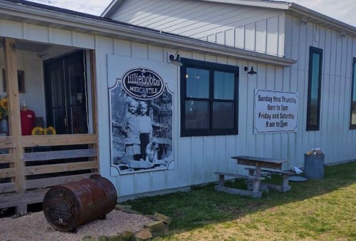 Exterior of a mercantile with a vintage photo on the wall, featuring a barrel and picnic table outside.