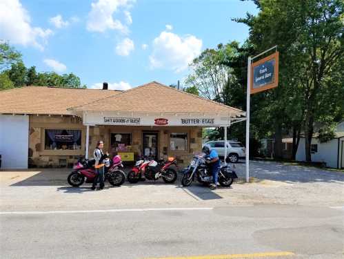 Two motorcyclists stand beside their bikes in front of a small store with a sign reading "Butter Eggs." Trees and blue sky in background.