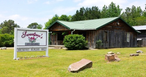 A rustic wooden building with a green metal roof, featuring a sign for "Sweetpea's" restaurant in a grassy area.