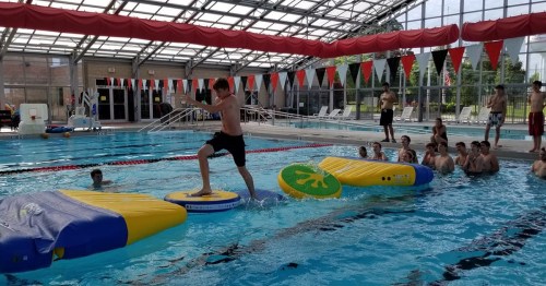 A shirtless boy balances on inflatable obstacles in a swimming pool, while others watch from the side.