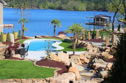 A serene lakeside view featuring a pool, landscaped garden, rocks, and palm trees under a clear blue sky.