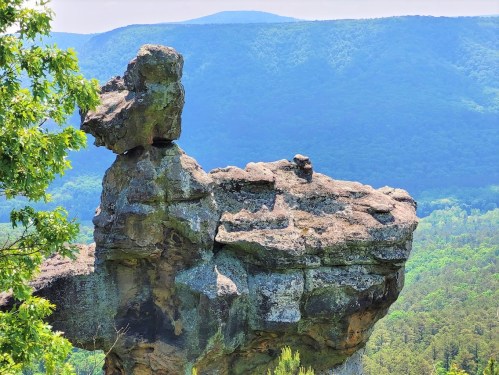 A large rock formation stands prominently against a backdrop of green mountains and a clear blue sky.