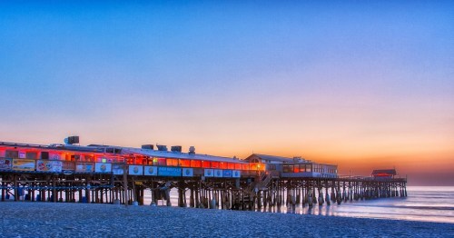 A colorful pier at sunset, with restaurants and shops illuminated against a serene ocean backdrop.