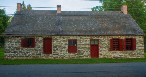 A historic stone house with a steep roof, red doors, and windows, set in a green landscape.