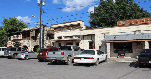 A row of rustic buildings with shops, parked cars, and power lines under a blue sky with scattered clouds.