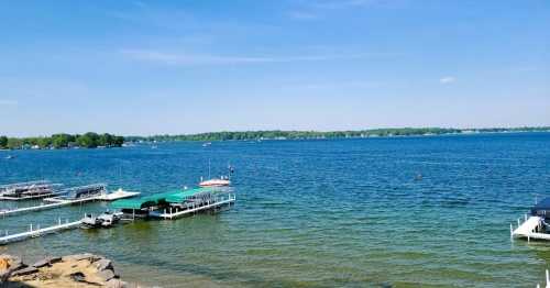 A serene lake view with boats docked, clear blue water, and a bright sky, surrounded by green trees.