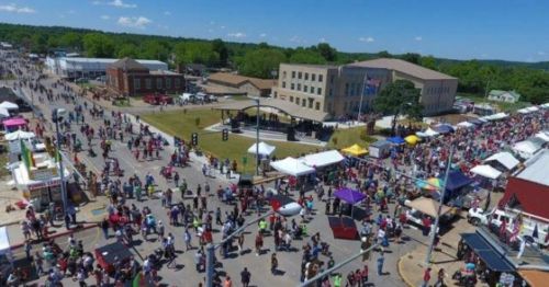 Aerial view of a bustling outdoor festival with crowds, food stalls, and a stage in a vibrant town setting.