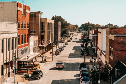 A sunny street view of a small town, featuring shops, parked cars, and trees lining the road.