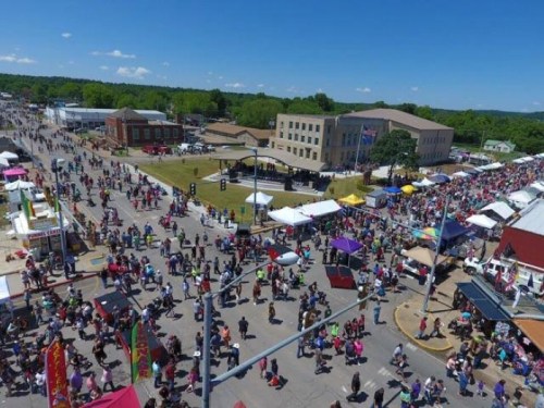 Aerial view of a bustling outdoor festival with crowds, food stalls, and colorful tents under a clear blue sky.