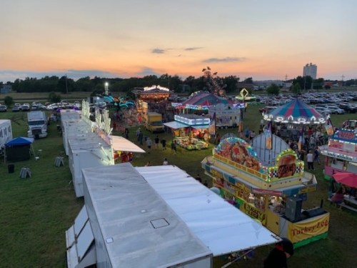 Aerial view of a vibrant fair at sunset, featuring colorful rides, food stalls, and a parking lot in the background.