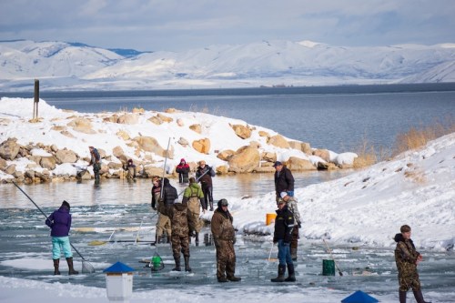 People ice fishing on a frozen lake surrounded by snow-covered mountains and rocky shores.