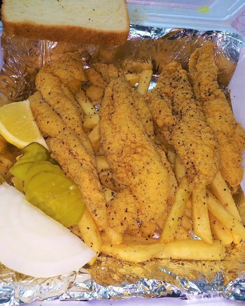 A serving of fried fish fillets with fries, pickles, onion, lemon wedges, and a slice of bread in a takeout container.