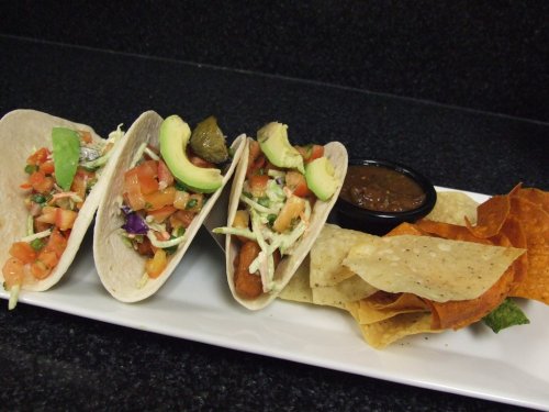 Three tacos filled with fresh toppings, served with a side of salsa and assorted tortilla chips on a white plate.