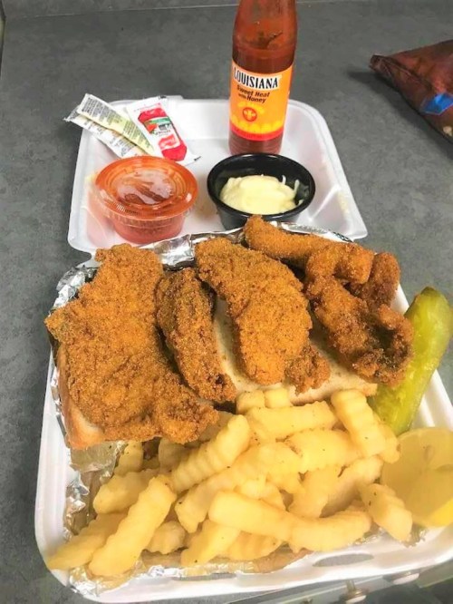 A plate of fried fish on bread, crinkle-cut fries, pickles, dipping sauces, and a drink.