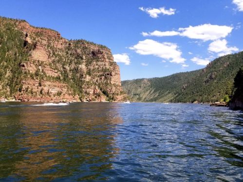 A serene river flows between towering cliffs and lush green hills under a clear blue sky with scattered clouds.