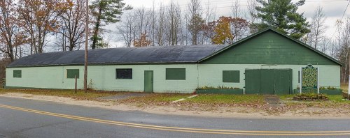 A long, green and white building with boarded windows, set against a backdrop of trees and a cloudy sky.