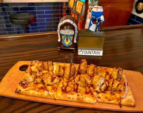 A flatbread pizza topped with chicken and drizzled sauce, next to a vintage jukebox figurine labeled "The Fountain."