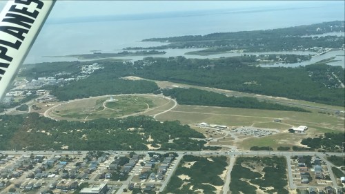 Aerial view of a coastal area with greenery, water, and a circular track, surrounded by residential buildings.