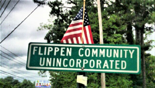 Sign for Flippen Community Unincorporated, with an American flag in the background and trees lining the street.
